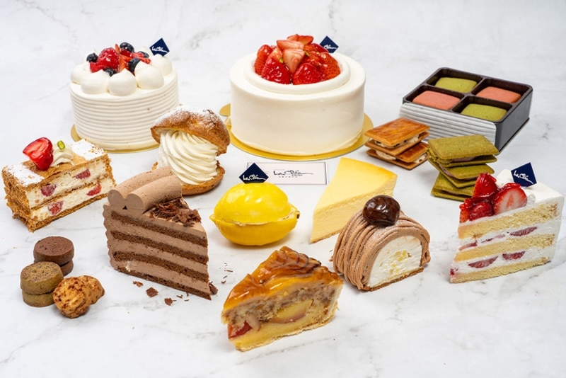 白い大理石調の台に、いちごのホールケーキ2台、モンブラン、チーズケーキ、レモン形のケーキ、チョコレートケーキ、ミルフィーユ、シュークリーム、抹茶のサンドクッキー、焼き菓子が並び、ケーキの上に紺色のLa cleaの小旗が立っている