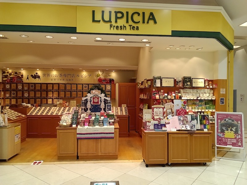 黄色い看板にLUPICIA FreshTEAと書かれた八王子オクトーレ1階のルピシア店舗。店内奥の壁一面に茶色い茶葉の箱が並び、手前の木目のカウンターに缶やギフト、白い案内板が置かれている