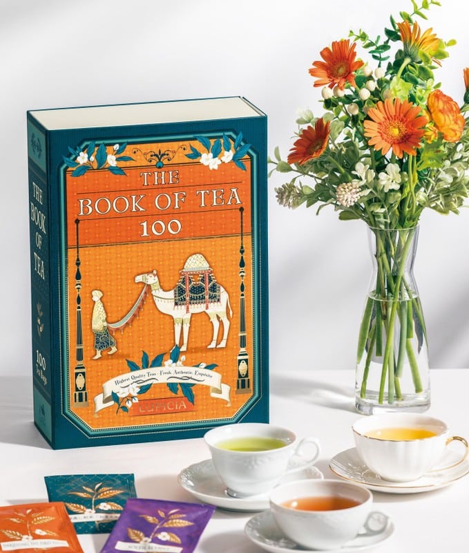 オレンジ色の表紙にラクダと人物が描かれたルピシアのギフト箱「THE BOOK OF TEA 100」。右にガーベラの花瓶、手前に緑茶と紅茶のカップ、紫とオレンジのティーバッグが置かれている