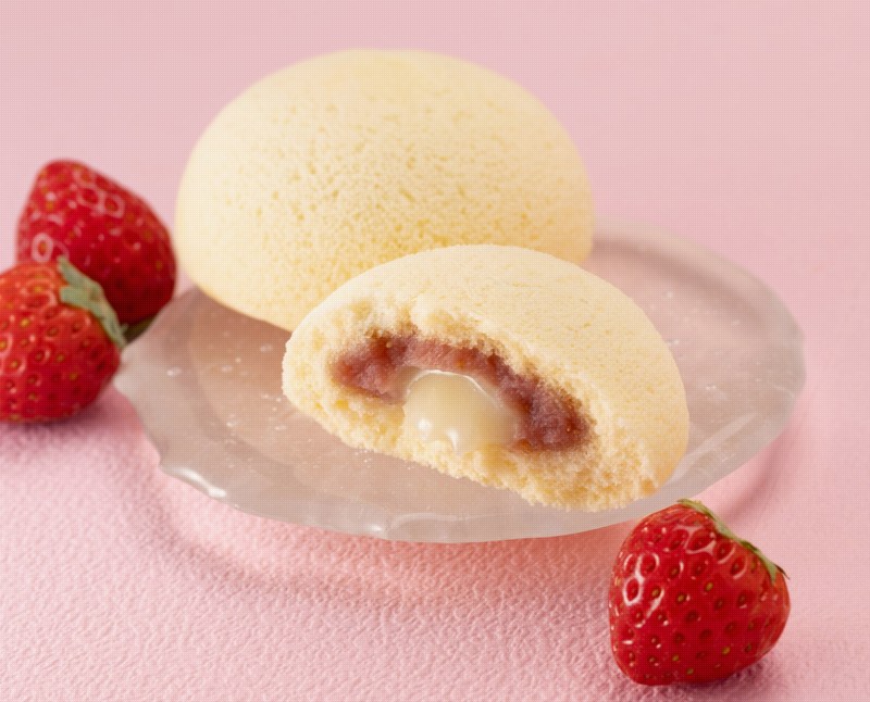ピンクの背景に、黄色い丸い蒸しケーキが2個。手前は半分に切られ、赤い苺カスタードと白い練乳の二層が見える。周りに赤い苺が3個