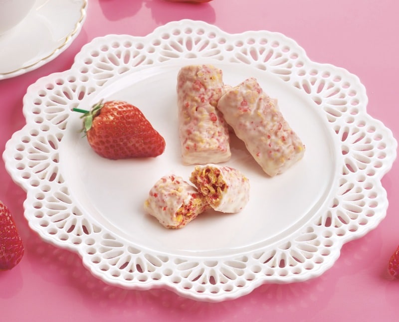 ピンクの背景に白いレース皿。白いチョコレートをまとった棒状のクランチが2本と、半分に割った断面、赤い苺が1個載っている