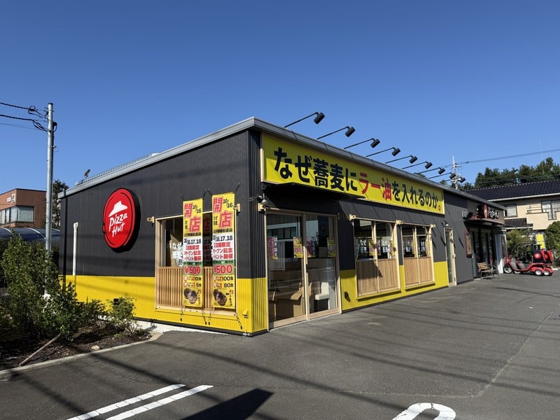 青空の下、左にピザハットの赤い屋根ロゴ、右になぜ蕎麦にラー油を入れるのか。の黄色い看板が並ぶ平屋。窓前に開店告知ののぼりが立ち、敷地は舗装された駐車場になっている
