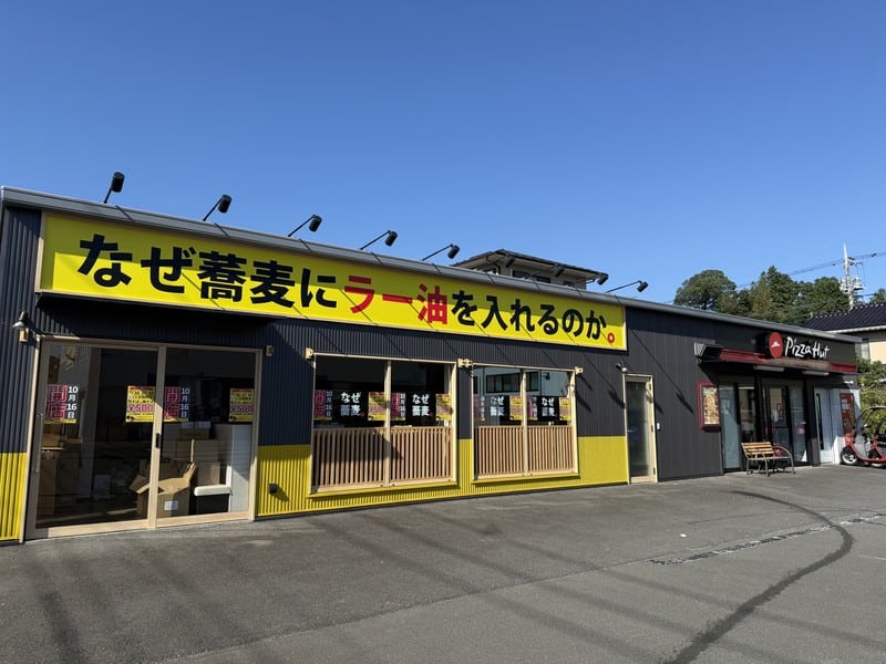 青空の下、黄色い看板になぜ蕎麦にラー油を入れるのか。と書かれた平屋店舗の正面。黒い壁と黄色い腰壁、木格子の窓、右端にピザハットの入口とベンチがある
