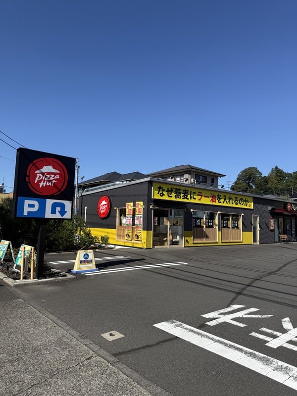 駐車場の白い矢印とピザハットのポール看板の奥に、なぜ蕎麦にラー油を入れるのか。の黄色い看板店舗が見える。敷地は広く舗装され、右側に白い停止線がある