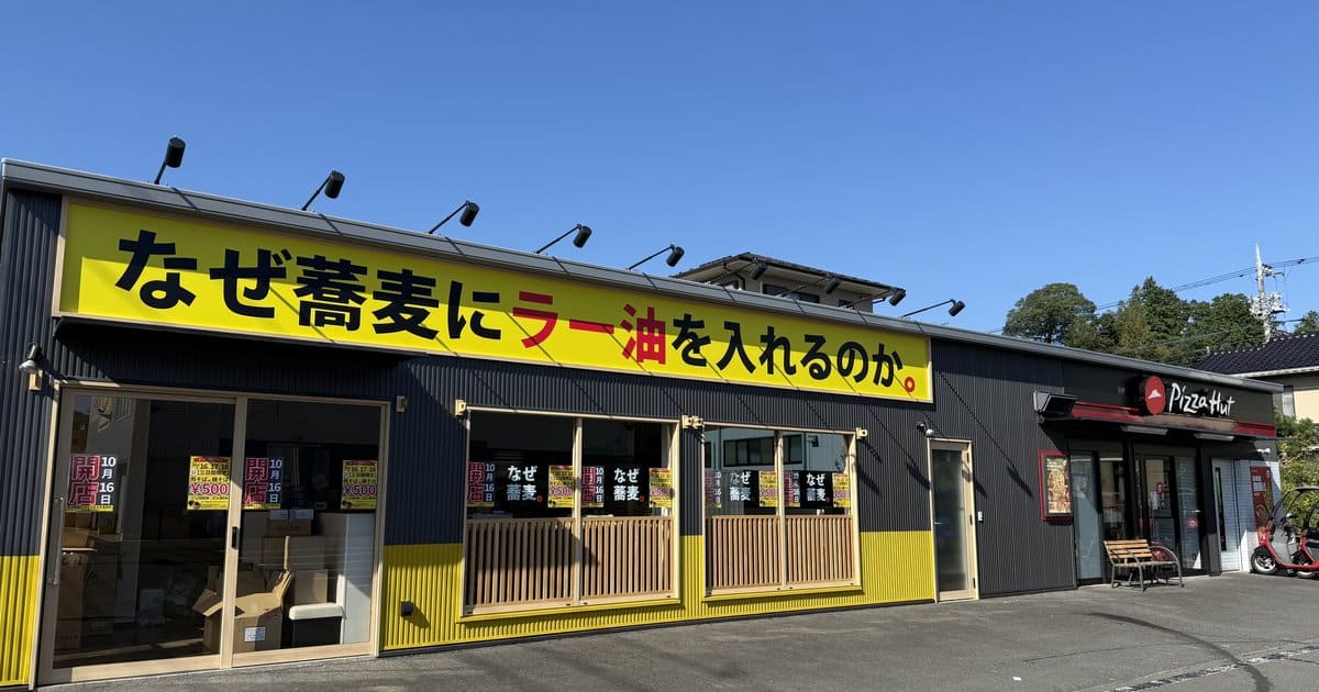 黄色い看板になぜ蕎麦にラー油を入れるのか。と書かれた平屋店舗の正面。右端にピザハットの入口がある