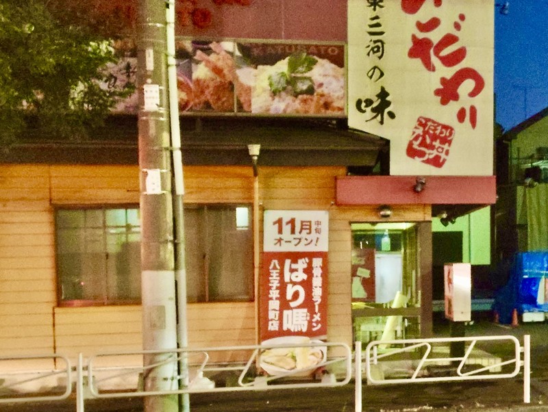 夜の木目外壁の店舗。上部にかつさとの三河の味の看板、中央に11月中旬オープンとばり馬八王子平岡町店の縦看板、手前に電柱と白い柵