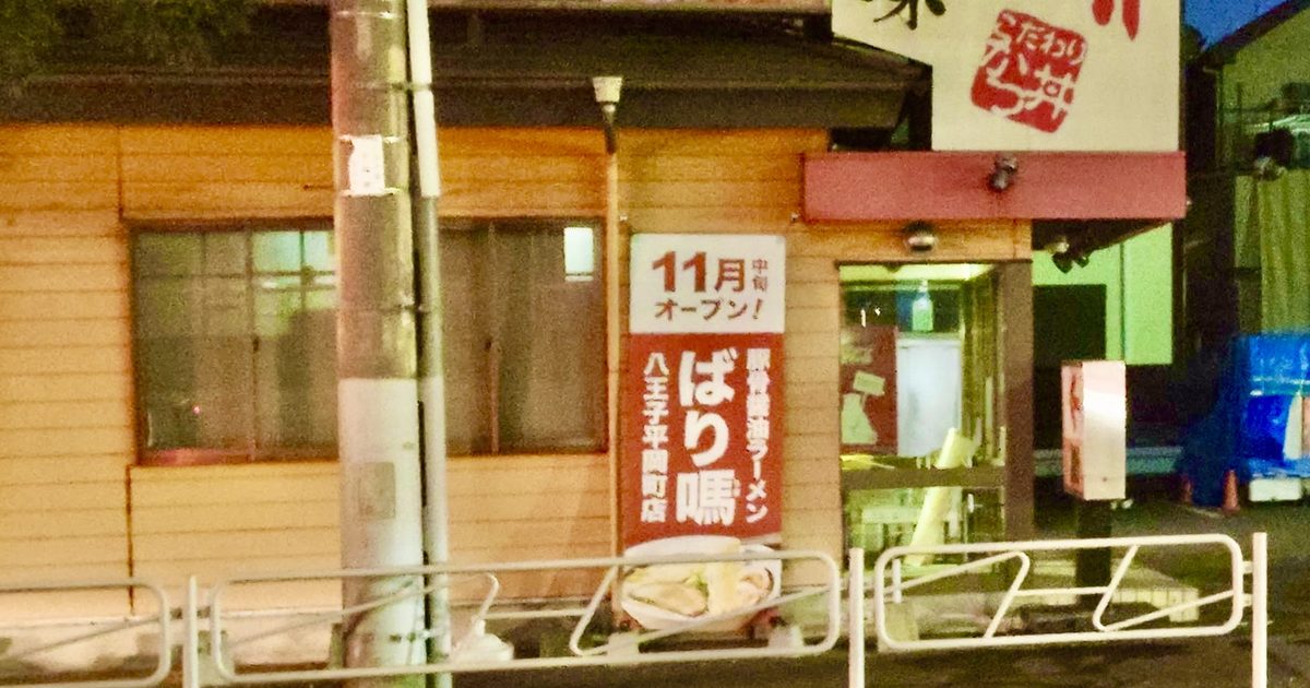 夜の木目外壁に11月中旬オープンとばり馬八王子平岡町店の縦看板が掲出され、手前に電柱と白い柵がある店舗外観