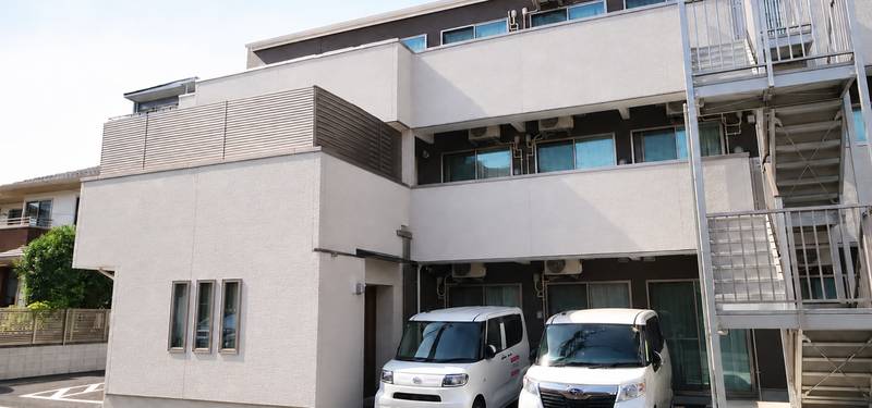 白い外壁の3階建て施設の外観。1階前に白いワゴン車が2台止まり、右側に鉄骨の避難階段がある