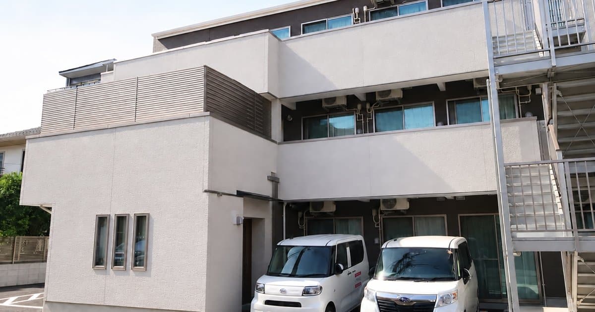 白い外壁の3階建て施設の外観。1階前に白いワゴン車が2台止まり、右側に鉄骨の避難階段がある
