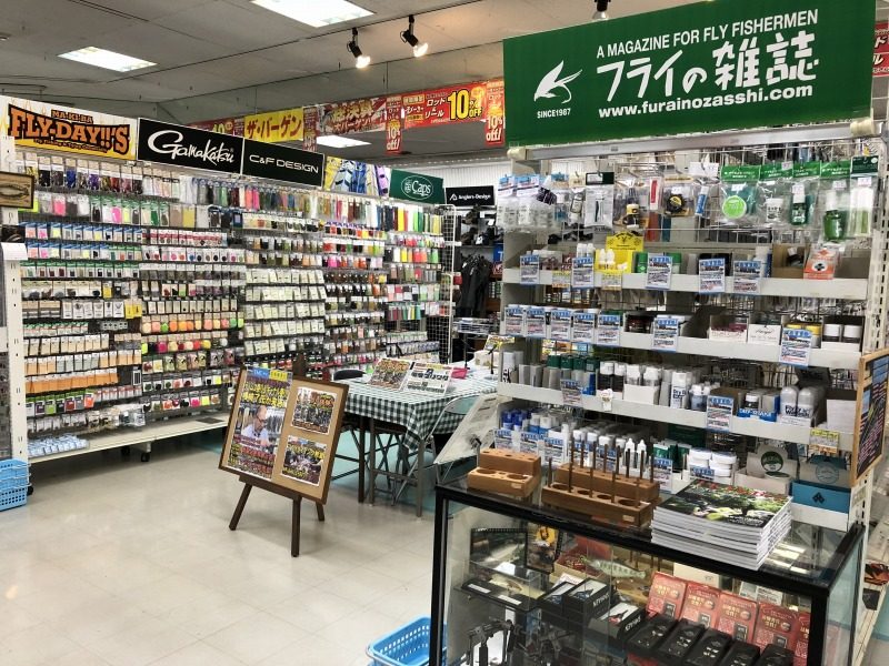 フライコーナーの店内。左に毛鉤や材料の棚、中央にチェックのテーブルと写真パネル、右にフライの雑誌の緑看板とタイイング用品の棚がある