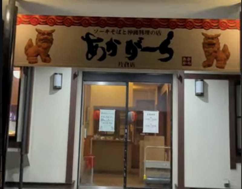 白い壁の店舗入口。上部の看板にソーキそばと沖縄料理の店、筆文字のあかがーら、片倉店、左右にシーサー。中央にガラスの自動ドアがあり、店内の照明が漏れている